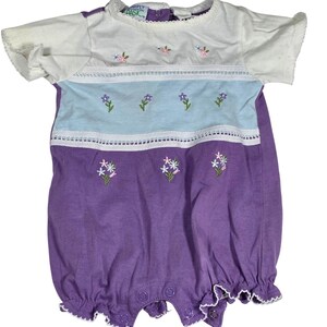 May include: A baby romper in white, light blue, and purple with floral embroidery. The top is white with short sleeves and a round neckline. The middle section is light blue, and the bottom is purple. The romper has snap closures and ruffled edges.