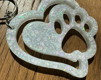 May include: A white, glittery keychain featuring a heart and paw print design. The keychain has a silver keyring attached. The heart shape surrounds the paw print, creating a unique and eye-catching accessory. The glitter adds a touch of sparkle.