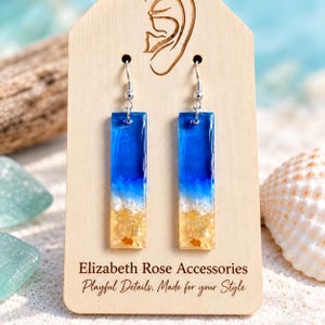 May include: A pair of rectangular drop earrings featuring a blue resin top transitioning into a white foam layer and a sandy base with embedded pebbles. The earrings are displayed on a light wood card labeled Elizabeth Rose Accessories.