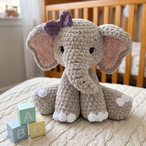 Puede incluir: Un peluche de elefante tejido a ganchillo en hilo marrón claro con orejas grandes que tienen secciones interiores rosas, un lazo morado en la cabeza y detalles blancos en los pies. Tres bloques de madera con las letras A, B y C están delante.