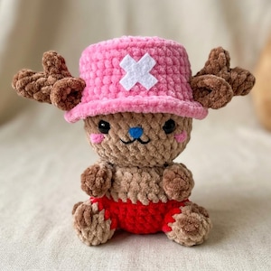 Könnte beinhalten: Eine gehäkelte Plüschpuppe mit beigem Körper, braunem Geweih und einem leuchtend rosa Hut mit weißem Kreuzemblem. Die Puppe hat schwarze Knopfaugen, eine kleine blaue Nase und trägt eine rote Hose. Die Textur ist durchgehend weich und flauschig.