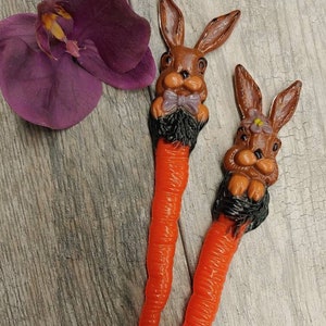 Puede incluir: Dos decoraciones de Pascua en forma de zanahoria naranja con cabezas de conejo. Los conejos tienen pelo marrón, ojos negros y una nariz rosa. Un conejo tiene una corbata de lazo morada y el otro una flor morada.