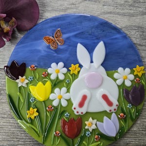 May include: A circular glass decoration with a blue sky backdrop and a green grassy foreground. It features a white bunny with a pink tail and red feet, surrounded by flowers and a butterfly.