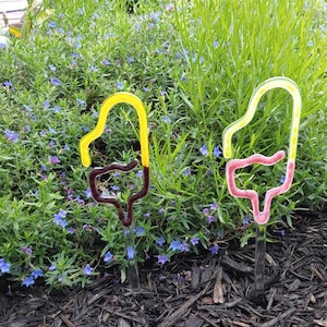 May include: Two glass garden stakes shaped like popsicles. The stake on the left is yellow and brown, and the stake on the right is pink and brown. The stakes are in a garden bed with mulch and blue flowers.