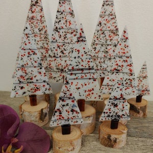 May include: A collection of decorative glass Christmas trees. The trees are white with red and black speckles, and sit on small wooden bases. The trees vary in size and shape, creating a festive display. A purple flower is in the foreground.