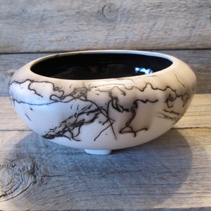 May include: A handmade, off-white ceramic bowl with a black interior. The exterior features a unique black lightning-like pattern. The bowl has a rounded shape and sits on three small feet, perfect for decorative use.
