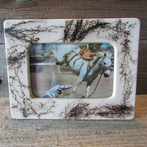 May include: Rectangular white picture frame with a rodeo scene. The frame has a decorative black squiggly design around the edges. The photo shows a bucking horse with a saddle and a cowboy in the arena.