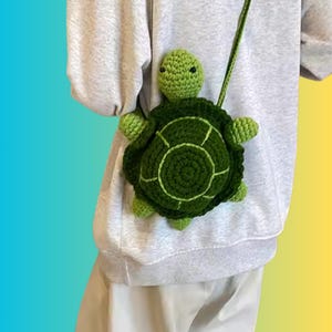 Puede incluir: Un bolso bandolera verde con forma de tortuga de ganchillo con una correa larga. El bolso presenta un diseño de caparazón detallado y una cabeza de tortuga. El bolso está hecho de hilo verde y tiene una apariencia caprichosa y hecha a mano.