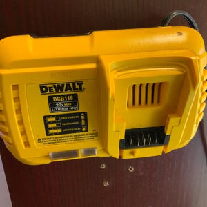 May include: A yellow DeWalt DCB118 20V Max Lithium Ion battery charger. The charger has a black power cord and features indicator lights for charging status. The DeWalt logo and model information are visible on the front.