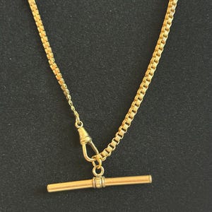 Puede incluir: Un collar dorado con un colgante de barra. La cadena es de estilo caja y el colgante tiene un diseño de barra en T. El collar se muestra sobre un fondo oscuro, resaltando su brillo metálico.
