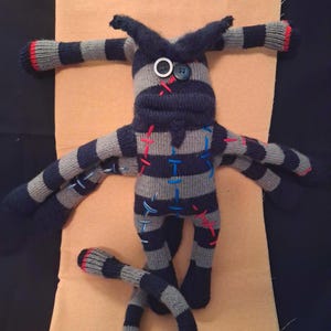 May include: A multi-limbed creature doll crafted from grey and navy blue striped knit fabric. It features two mismatched button eyes, one white and one dark blue, with red stitched detailing. The doll has six arms, two antennae-like appendages, and a long striped tail, all accented with red and blue decorative stitching throughout the body.
