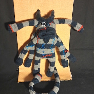 May include: Handmade sock monster doll in gray and navy blue stripes with red and blue stitching details. The doll has horns, a button eye, and a long, curved tail. A unique, whimsical stuffed animal.