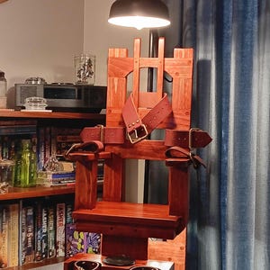 Może przedstawiać: Drewniana replika krzesła elektrycznego ze skórzanymi paskami i lampą nad nim. Krzesło jest zabarwione na ciemny, czerwonawy brąz i ma małą platformę u dołu.