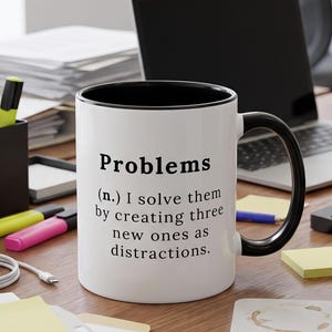 Può includere: Una tazza in ceramica bianca con interno e manico neri, caratterizzata dal testo stampato in nero: Problems (n.) I solve them by creating three new ones as distractions. La tazza è cilindrica con una finitura liscia e un manico laterale.
