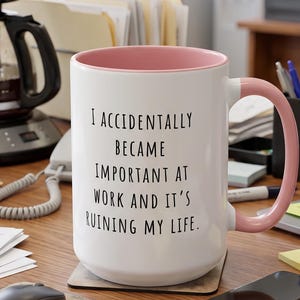 Puede incluir: Una taza de cerámica blanca con interior y asa de color rosa suave. La taza presenta un texto impreso en negro que dice: I ACCIDENTALLY BECAME IMPORTANT AT WORK AND IT'S RUINING MY LIFE. La taza tiene una forma cilíndrica y un asa lateral.