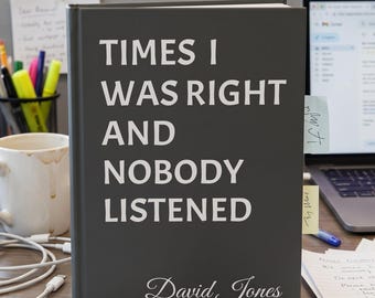 May include: A dark gray hardcover book with white text that reads "TIMES I WAS RIGHT AND NOBODY LISTENED" and "David Jones" in a script font. The book is positioned on a desk with office supplies, a white mug, and a laptop in the background.