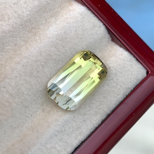 Könnte beinhalten: Ein rechteckiger, facettierter Edelstein mit einem blassgelben und hellgrünen Farbton. Der Stein hat ein klares, transparentes Aussehen mit scharfen, geometrischen Facetten, die das Licht reflektieren und einen hellen, leuchtenden Effekt erzeugen.