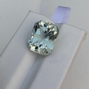 Peut inclure: Une pierre pr&eacute;cieuse aigue-marine rectangulaire et facett&eacute;e, de couleur bleu-vert p&acirc;le. La gemme est pos&eacute;e sur une surface blanche avec un ruban blanc en arri&egrave;re-plan. Les facettes de la pierre pr&eacute;cieuse refl&egrave;tent la lumi&egrave;re.