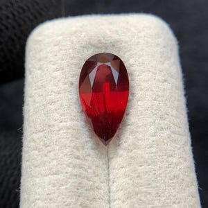 Könnte beinhalten: Ein birnenförmiger, facettierter roter Edelstein mit tiefroter Farbe und spitzem Ende. Der Stein weist einen symmetrischen Schliff mit mehreren dreieckigen und drachenförmigen Facetten auf, die das Licht reflektieren und ein klares Aussehen erzeugen.