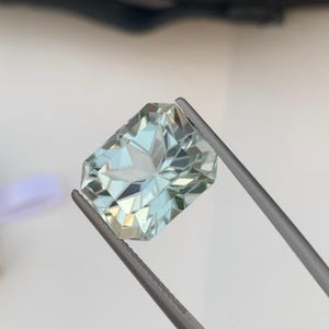 Peut inclure: Une pierre pr&eacute;cieuse rectangulaire bleu clair tenue par une pince. La gemme est taill&eacute;e &agrave; facettes, refl&eacute;tant la lumi&egrave;re et cr&eacute;ant un motif en &eacute;toile au centre. La couleur de la pierre est un bleu p&acirc;le translucide, avec des reflets blancs.
