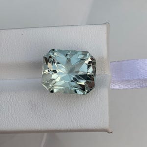 Peut inclure: Une pierre pr&eacute;cieuse rectangulaire bleu clair avec une taille &agrave; facettes. La gemme est pos&eacute;e sur une surface blanche avec un ruban blanc visible en arri&egrave;re-plan. Les facettes de la pierre pr&eacute;cieuse refl&egrave;tent la lumi&egrave;re.