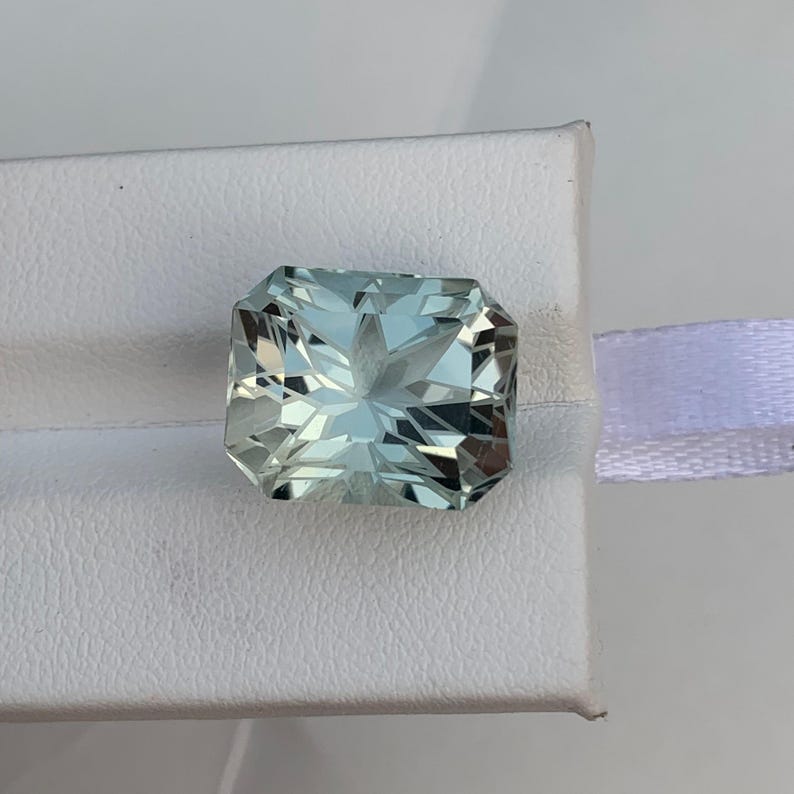 Peut inclure: Une pierre pr&eacute;cieuse rectangulaire bleu clair avec une taille &agrave; facettes. La gemme est pos&eacute;e sur une surface blanche avec un ruban blanc visible en arri&egrave;re-plan. Les facettes de la pierre pr&eacute;cieuse refl&egrave;tent la lumi&egrave;re.