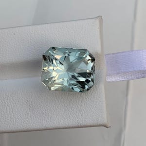Peut inclure: Une pierre pr&eacute;cieuse rectangulaire bleu clair avec de multiples facettes, sur fond blanc. La gemme a un aspect clair et cristallin, refl&eacute;tant la lumi&egrave;re. La taille de la pierre est visible, mettant en valeur ses angles et ses plans. La gemme est probablement de l'aigue-marine.
