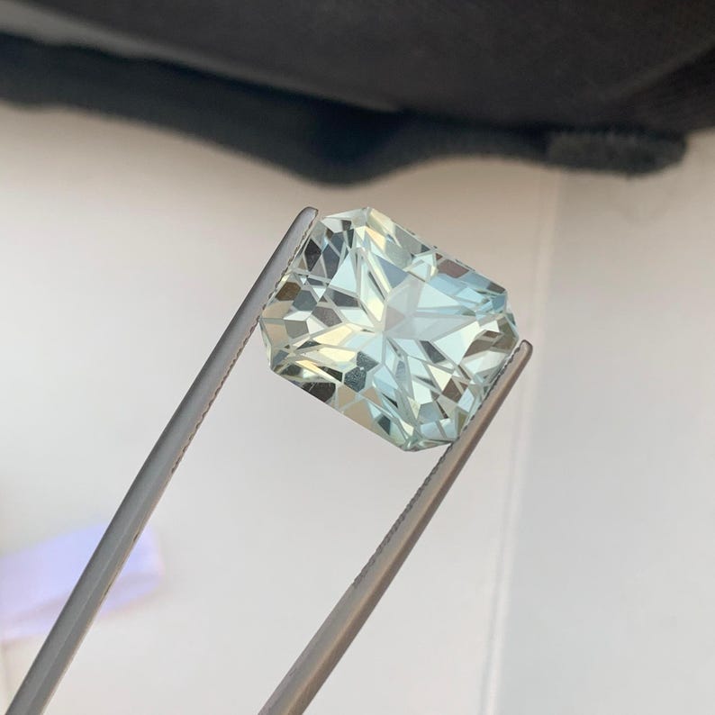 Peut inclure: Une pierre pr&eacute;cieuse rectangulaire bleu clair, tenue par une pince argent&eacute;e. La gemme est taill&eacute;e &agrave; facettes, refl&eacute;tant la lumi&egrave;re et r&eacute;v&eacute;lant sa structure interne. La couleur de la gemme est un bleu clair translucide.