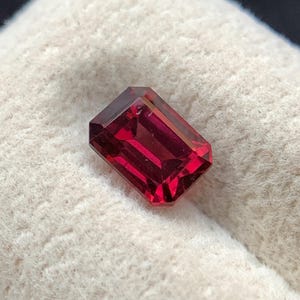 May include: A faceted, emerald-cut gemstone with a deep, vibrant red color. The stone features sharp, clean edges and a polished surface that reflects light, showcasing its transparent quality and geometric, rectangular shape.