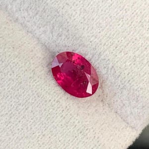 May include: A faceted, oval-shaped ruby gemstone. The gem is a vibrant red color, with visible internal inclusions. The stone is set against a white, textured background, highlighting its color and clarity. This gemstone is suitable for jewelry making.