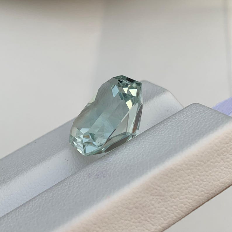 Peut inclure: Une pierre pr&eacute;cieuse bleu clair taill&eacute;e &agrave; facettes. La gemme pr&eacute;sente de multiples facettes qui r&eacute;fl&eacute;chissent la lumi&egrave;re, cr&eacute;ant un effet scintillant. La pierre est pos&eacute;e sur un fond blanc. Environ 2 cm de long.