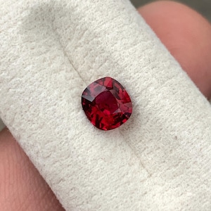 Könnte beinhalten: Ein Edelstein im Kissenschliff mit einer tiefen, gesättigten roten Farbe und zahlreichen Facetten, die das Licht reflektieren. Der Stein liegt auf einer strukturierten, cremeweißen Stoffoberfläche und zeigt seine klare, transparente Beschaffenheit.