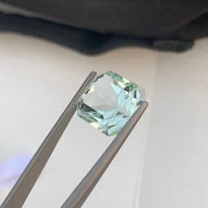 Peut inclure: Une pierre pr&eacute;cieuse bleu clair, taill&eacute;e en forme carr&eacute;e, tenue par une pince argent&eacute;e. La gemme pr&eacute;sente de multiples facettes qui r&eacute;fl&eacute;chissent la lumi&egrave;re. La pierre est d'un bleu clair translucide.