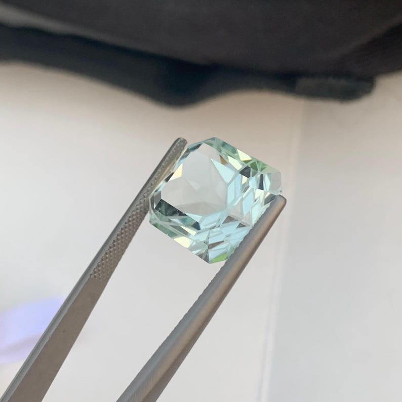 Peut inclure: Une pierre pr&eacute;cieuse bleu clair, taill&eacute;e en forme carr&eacute;e, tenue par une pince argent&eacute;e. La gemme pr&eacute;sente de multiples facettes qui r&eacute;fl&eacute;chissent la lumi&egrave;re. La pierre est d'un bleu clair translucide.
