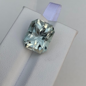 Peut inclure: Une pierre pr&eacute;cieuse aigue-marine rectangulaire et facett&eacute;e, de couleur bleu-vert p&acirc;le. La pierre est pos&eacute;e sur un fond blanc, mettant en valeur ses motifs internes complexes et ses qualit&eacute;s r&eacute;fl&eacute;chissantes. La taille de la pierre cr&eacute;e un effet d'&eacute;clat.