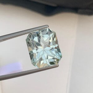 Peut inclure: Une pierre pr&eacute;cieuse rectangulaire bleu clair tenue par une pince. La gemme pr&eacute;sente une taille complexe &agrave; multiples facettes, refl&eacute;tant la lumi&egrave;re et cr&eacute;ant un motif en &eacute;toile au centre. La couleur de la pierre est un bleu p&acirc;le et translucide.