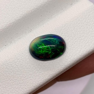 May include: Oval-shaped black opal gemstone with vibrant green, blue, and red internal colors. The polished stone displays a play of color, set against a dark background. The opal is displayed on a white surface.
