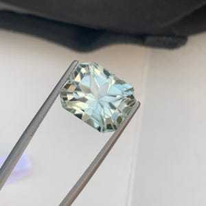 Peut inclure: Une pierre pr&eacute;cieuse bleu clair &agrave; facettes, tenue par une pince argent&eacute;e. La gemme a une forme rectangulaire avec de multiples facettes qui r&eacute;fl&eacute;chissent la lumi&egrave;re, cr&eacute;ant un effet scintillant. La couleur de la pierre est bleu p&acirc;le et froid.