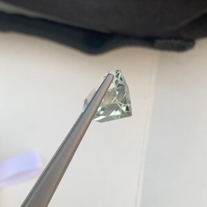Peut inclure: Une pierre pr&eacute;cieuse verte p&acirc;le, taill&eacute;e en forme triangulaire, est maintenue par une pince argent&eacute;e. Ses multiples facettes r&eacute;fl&eacute;chissent la lumi&egrave;re. Le fond est blanc, avec une ombre sombre en haut.