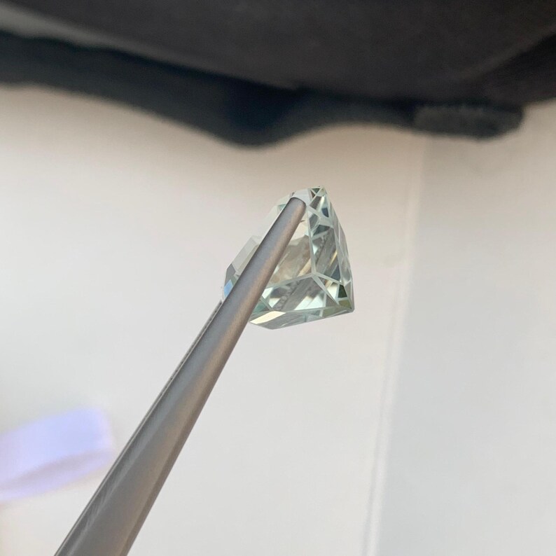 Peut inclure: Une pierre pr&eacute;cieuse verte p&acirc;le, taill&eacute;e en forme triangulaire, est maintenue par une pince argent&eacute;e. Ses multiples facettes r&eacute;fl&eacute;chissent la lumi&egrave;re. Le fond est blanc, avec une ombre sombre en haut.
