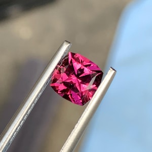 Puede incluir: Una gema de color magenta con corte tipo cojín, sostenida firmemente por unas pinzas de metal plateado. La piedra presenta una superficie compleja con múltiples facetas que reflejan la luz, creando destellos en rosa intenso y rojo.