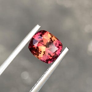 Puede incluir: Una piedra preciosa de corte cojín que muestra un degradado vibrante de tonos rosa intenso, magenta y naranja cálido. La piedra presenta una superficie facetada que refleja la luz, sostenida por dos puntas metálicas de pinzas plateadas.