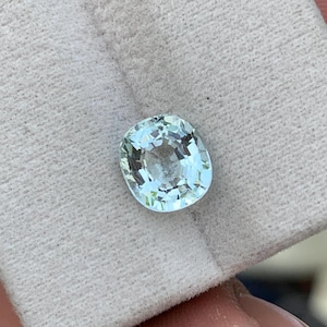 Op de afbeelding: Een lichtblauwe, ovaal geslepen edelsteen met een gefacetteerd oppervlak. De edelsteen ligt op een witte stof, waardoor de helderheid en kleur worden benadrukt. De facetten van de edelsteen reflecteren licht, wat een sprankelend effect creëert. Het thema is sieraden en edelstenen.