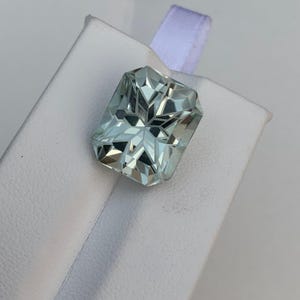 Peut inclure: Une pierre pr&eacute;cieuse rectangulaire &agrave; facettes, de couleur bleu-vert p&acirc;le. La gemme est pos&eacute;e sur une surface blanche avec un ruban blanc. Les facettes cr&eacute;ent un effet scintillant, refl&eacute;tant la lumi&egrave;re. Cette pierre est probablement pour la joaillerie.