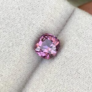Puede incluir: Una gema facetada, de talla cojín, en tonos rosa y púrpura. La gema se sostiene sobre una tela blanca texturizada. Las facetas de la piedra reflejan la luz, creando un efecto brillante. Esta gema es para joyería.