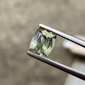 May include: A cushion-cut gemstone with a pale olive-green hue and visible internal inclusions. The faceted stone displays a translucent quality with light reflecting off its polished surfaces, held securely by a pair of serrated metal tweezers.