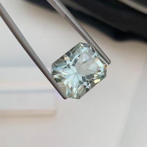 Peut inclure: Une pierre pr&eacute;cieuse rectangulaire bleu-vert clair tenue par une pince. La gemme a une taille complexe &agrave; multiples facettes, refl&eacute;tant la lumi&egrave;re. La couleur de la gemme est bleu-vert p&acirc;le et translucide, avec un aspect brillant et &eacute;tincelant.