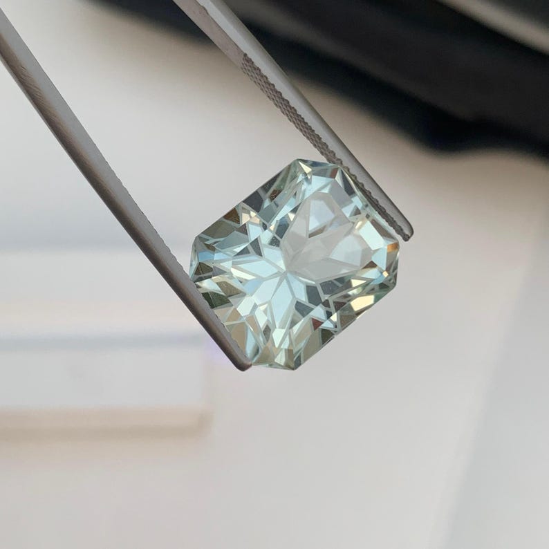Peut inclure: Une pierre pr&eacute;cieuse rectangulaire bleu-vert clair tenue par une pince. La gemme a une taille complexe &agrave; multiples facettes, refl&eacute;tant la lumi&egrave;re. La couleur de la gemme est bleu-vert p&acirc;le et translucide, avec un aspect brillant et &eacute;tincelant.