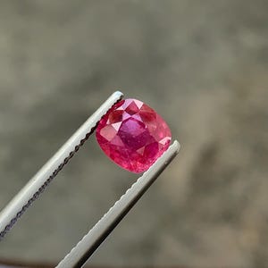 Könnte beinhalten: Ein facettierter, kissenförmiger Rubin, der von einer Pinzette gehalten wird. Der Rubin zeigt einen tiefroten Farbton mit inneren Facetten, die das Licht reflektieren. Die Pinzette ist silberfarben, und der Hintergrund ist ein verschwommenes Grau.