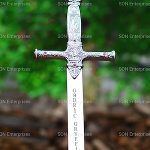 May include: A silver-toned metal sword featuring an ornate hilt with red gemstone accents. The blade is engraved with the vertical text GODRIC GRYFFINDOR. The crossguard and pommel display intricate relief patterns and small red circular inlays.